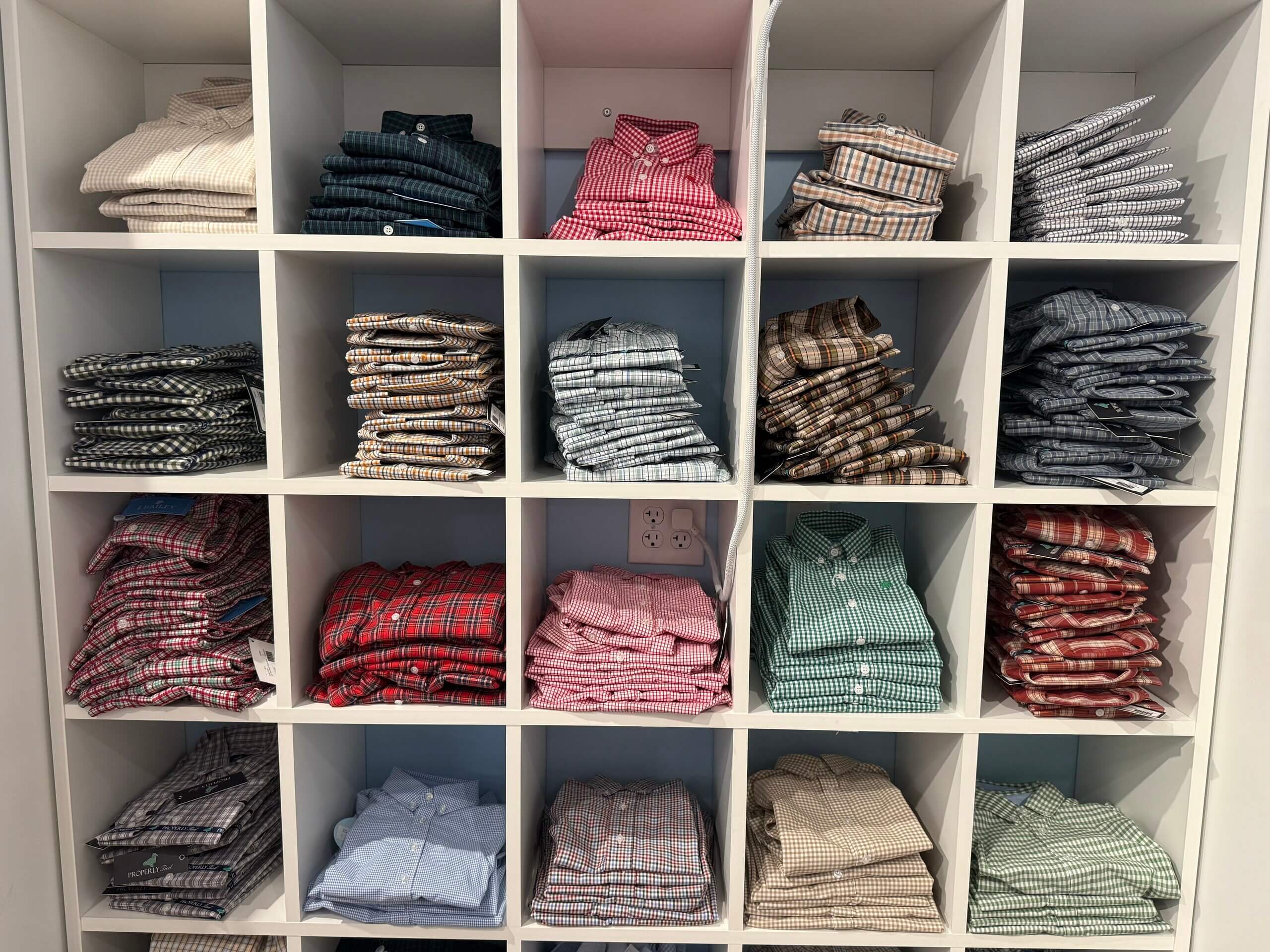 A shelving unit with sixteen cubbies, each holding neatly folded long-sleeve button-up plaid and solid shirts in various colors and patterns, creates a display as organized and vibrant as a Lili Pad in full bloom.