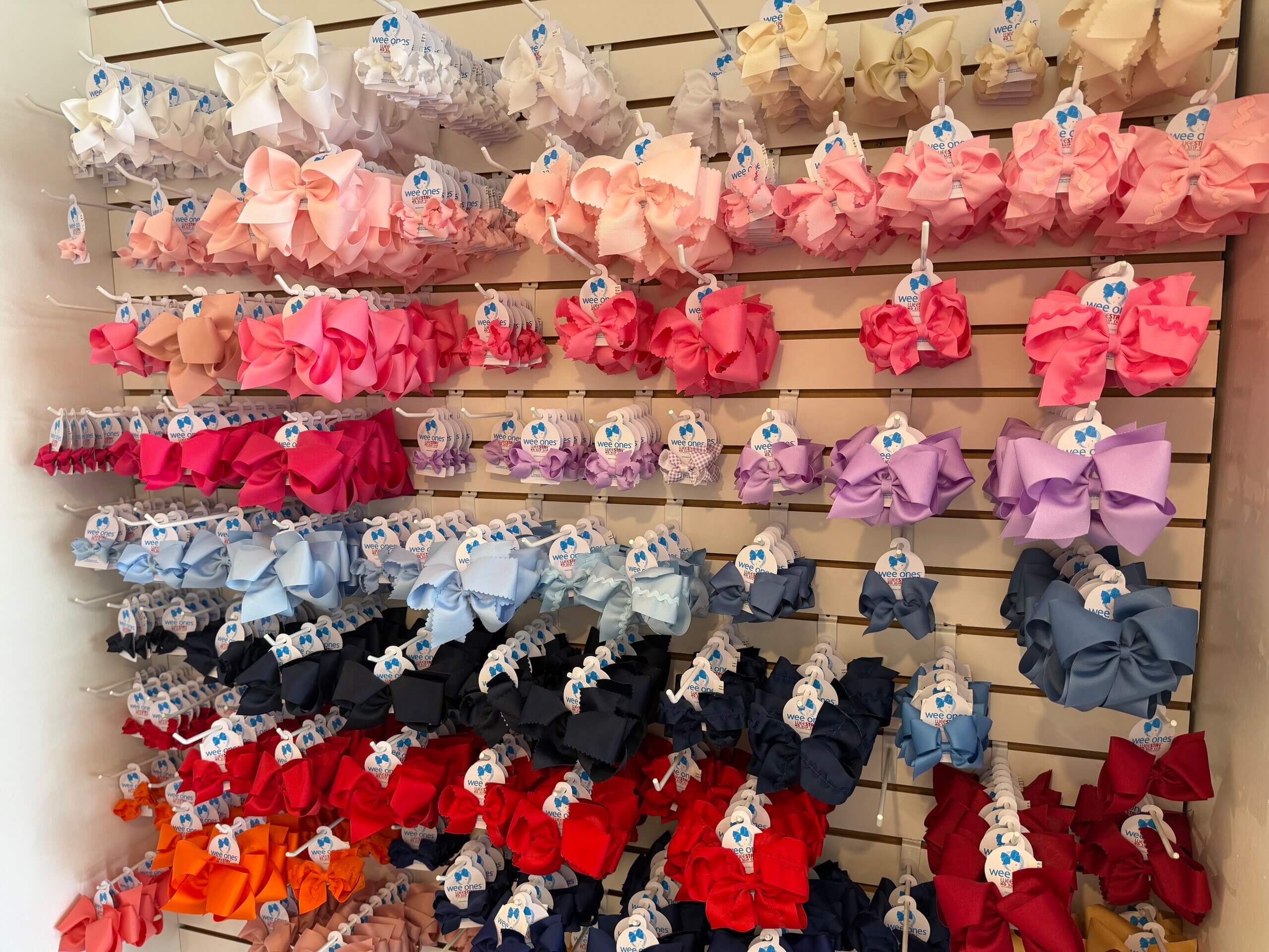 A display wall filled with hair bows organized by color, ranging from pinks and reds to blues and blacks, each Lili Pad bow hanging on a hook with a price tag.