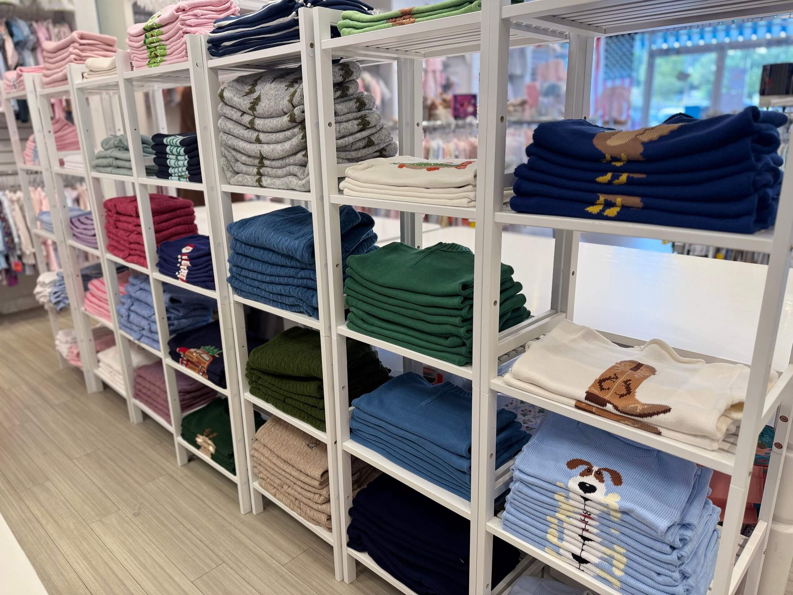 Shelves of neatly folded sweaters in various colors and designs, including playful animal and boot motifs, are displayed in a well-lit clothing store featuring the charming Lili Pad collection.