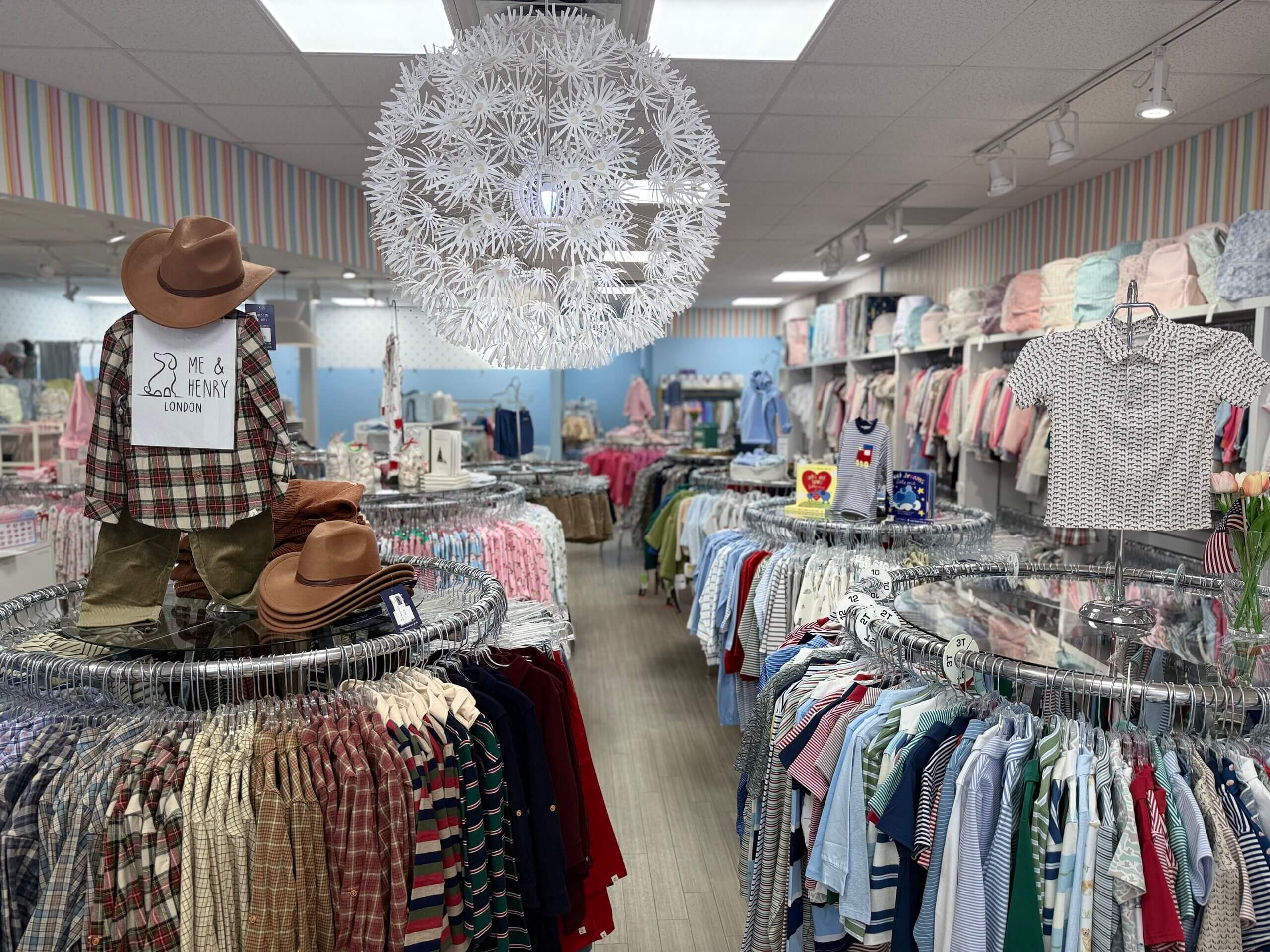 Children’s clothing store, Lili Pad, with racks of colorful shirts and outfits, hats on display, and a large decorative white ceiling light fixture in the center.