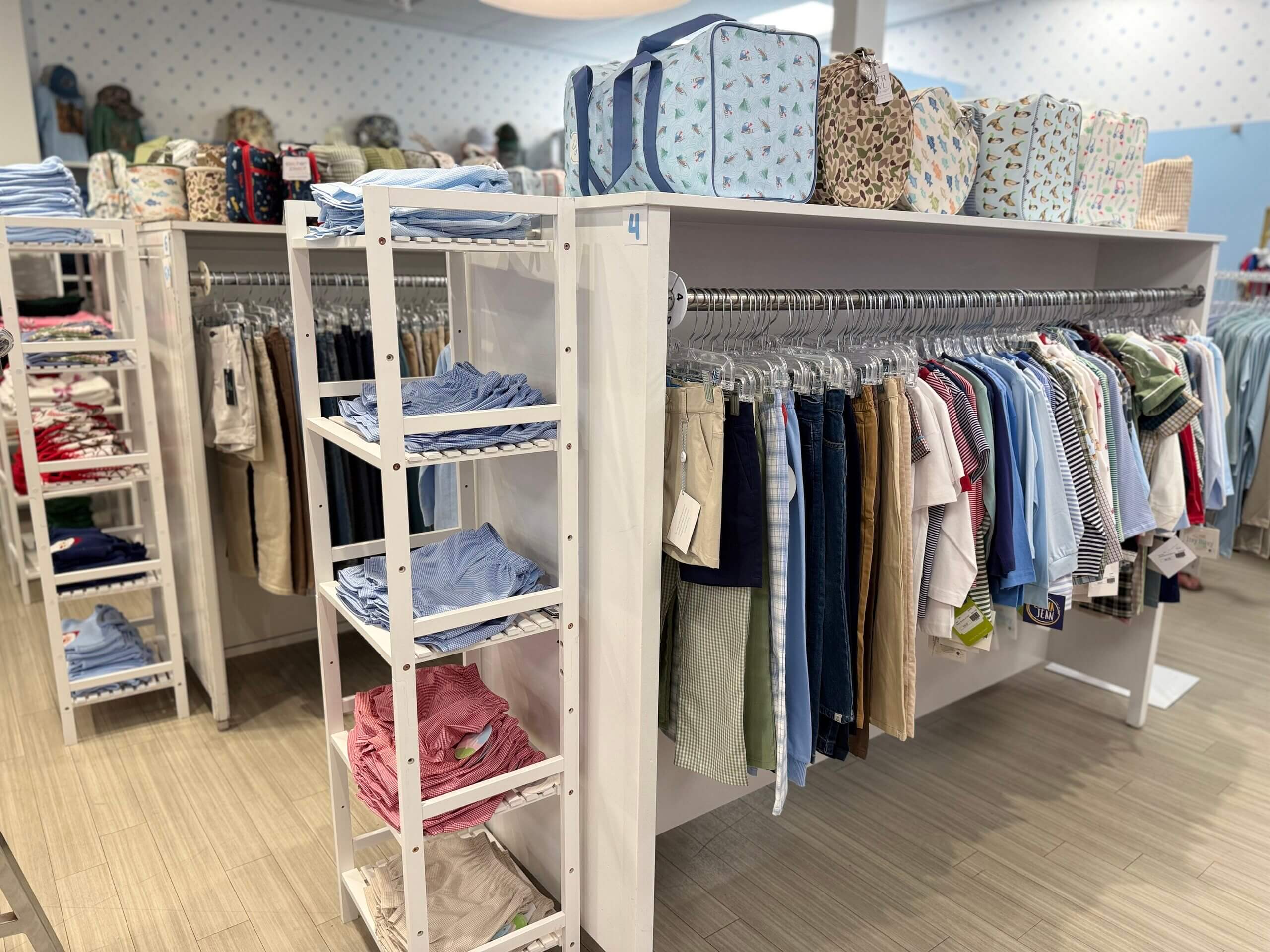 Clothing store display with folded shirts, shorts, and hanging shirts and pants. Patterned bags, including the stylish Lili Pad collection, are arranged on the top shelf. The floor and furniture are light-colored.