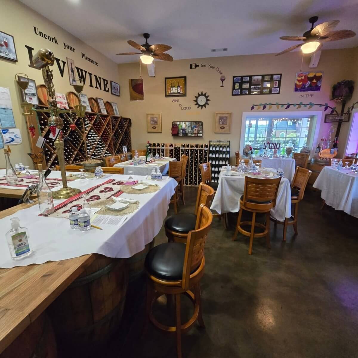 Wine tasting room with set tables, wooden chairs, wine racks, framed wall art, ceiling fans, and a window—perfect for those exploring Birmingham wine tours or seeking the charm of Birmingham wineries.