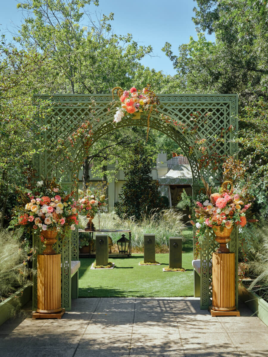 A green lattice arch decorated with pink and orange flowers stands in a garden, flanked by two large floral arrangements on wooden pedestals, with greenery and trees in the background—creating an enchanting setting reminiscent of Antiques at the Garden, celebrating 20 years of elegance in Birmingham.