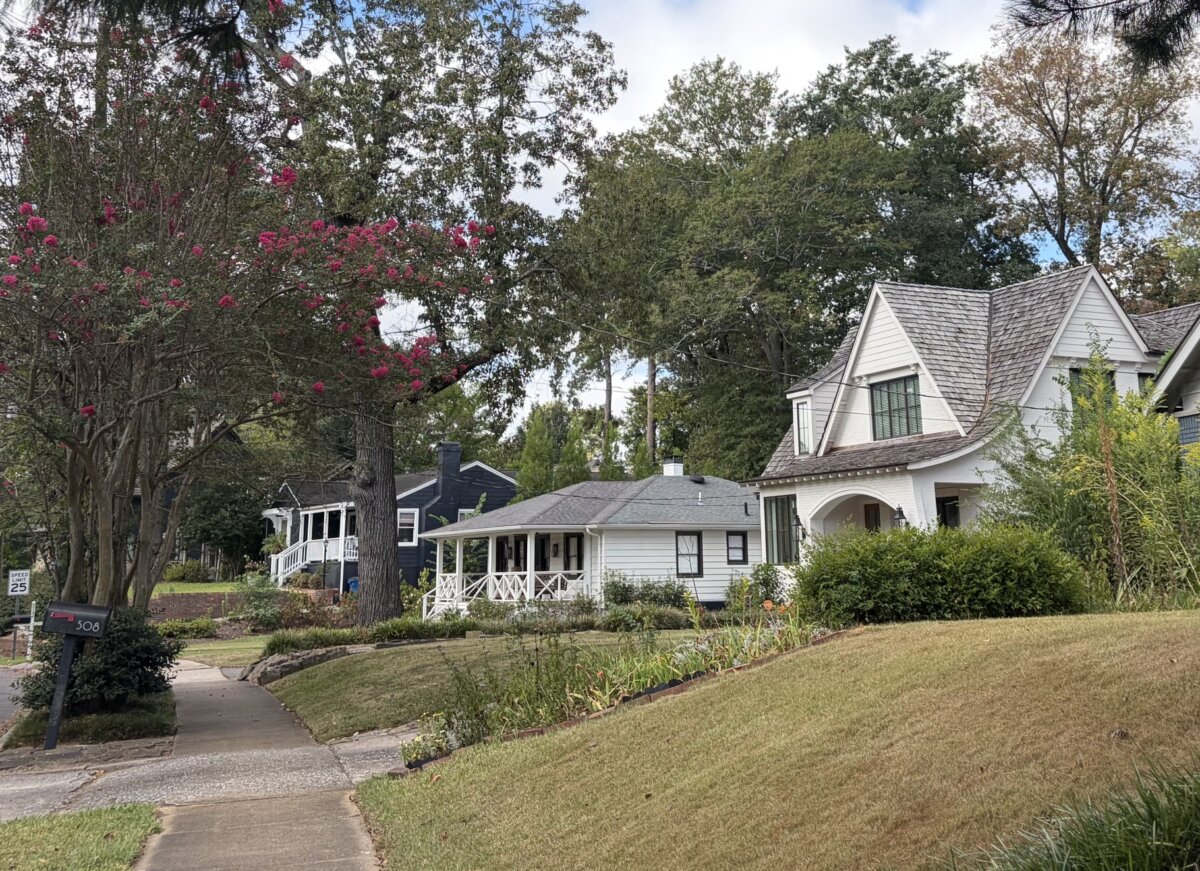 5 of Birmingham's Most Charming Walks - 7 A quiet suburban street with several houses, green lawns, blooming pink flowers on a tree, and a sidewalk curving through the neighborhood on a partly cloudy day—perfect for charming walks or those who enjoy exploring Birmingham walking trails.