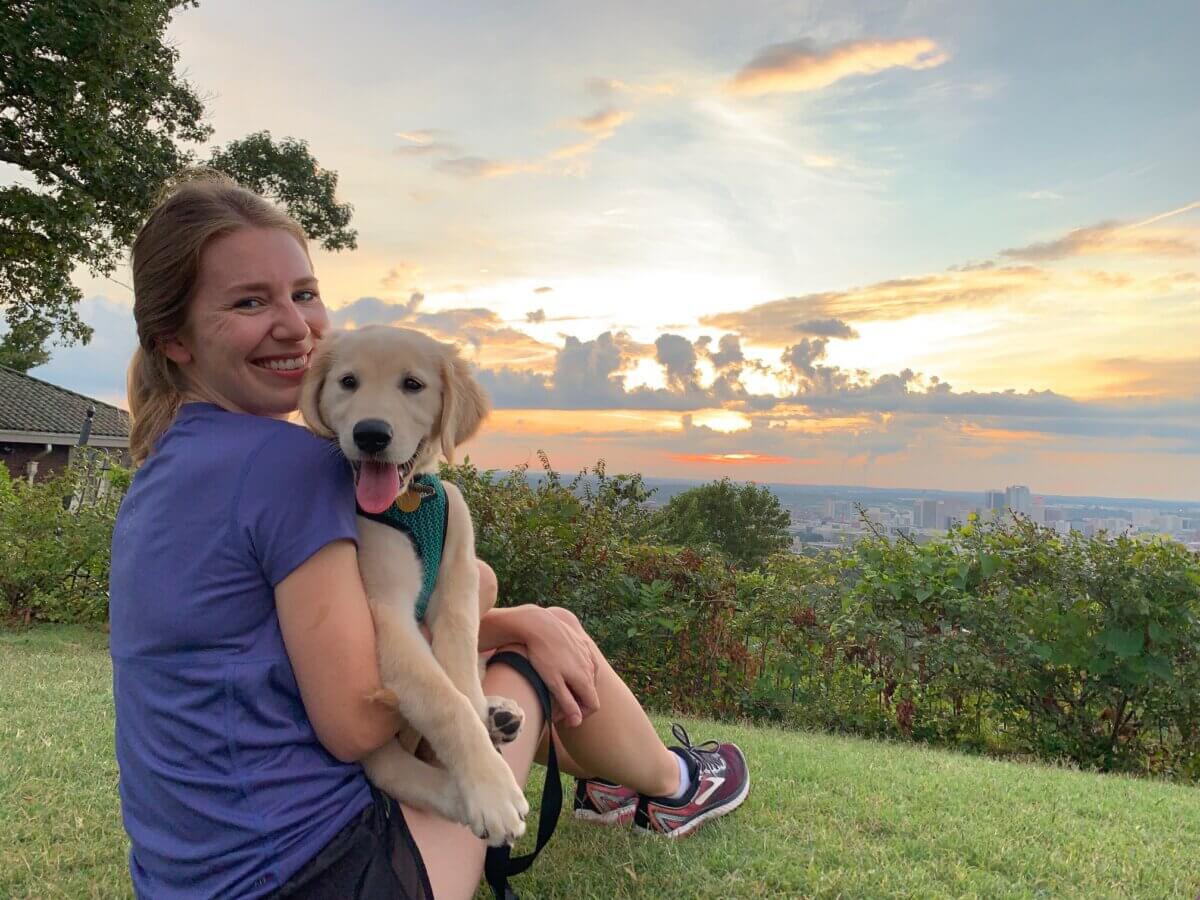 5 of Birmingham's Most Charming Walks - 2 A woman sits on grass at sunset, smiling and holding a golden retriever puppy after enjoying one of the charming Birmingham walks. A cityscape and clouds are visible in the background.