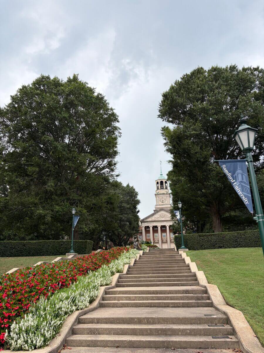 5 of Birmingham's Most Charming Walks - 4 Wide stone steps lead up through red and white flower beds to a building with a tall clock tower, framed by trees under a cloudy sky. A campus banner hangs from a lamp post on the right, marking this spot as one of the most charming walks along popular Birmingham walking routes.
