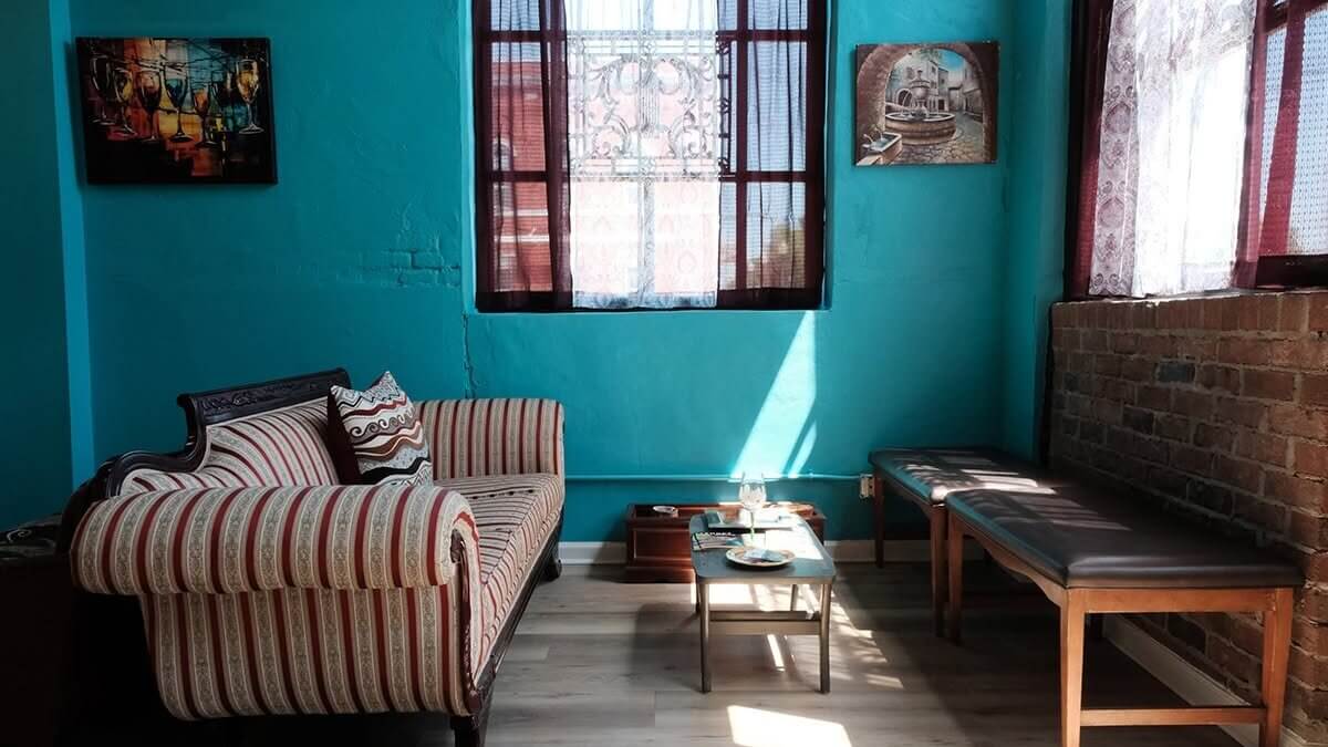 A cozy room with teal walls, a striped sofa, a bench, a small coffee table, wall art, and sunlight streaming through curtained windows onto a wooden floor—a perfect spot to unwind after touring the gorgeous wineries near Birmingham.