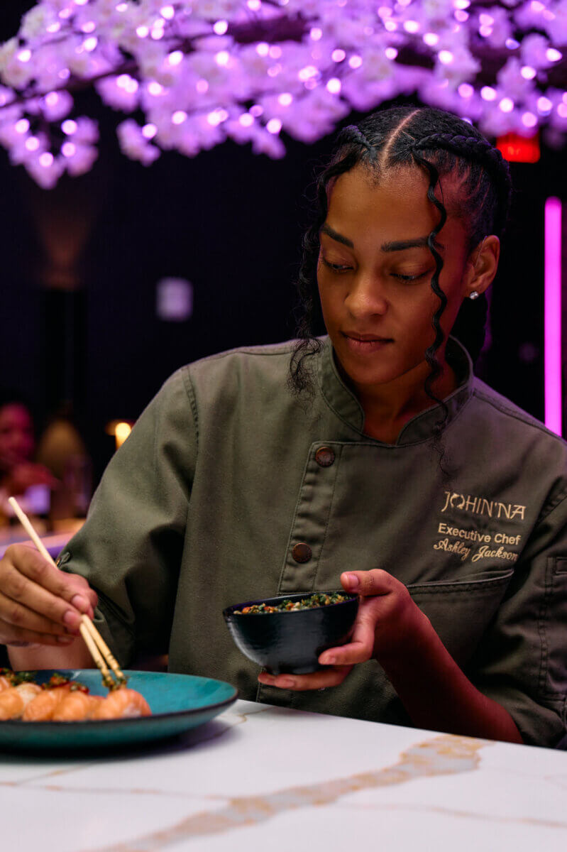 Southern Roots, Japanese Technique: Inside Chef Ashley Jackson's Johin'na - 3 Chef Ashley in a green uniform uses chopsticks to add garnish to a dish, with a bowl in hand, under purple decorative lighting in Birmingham.