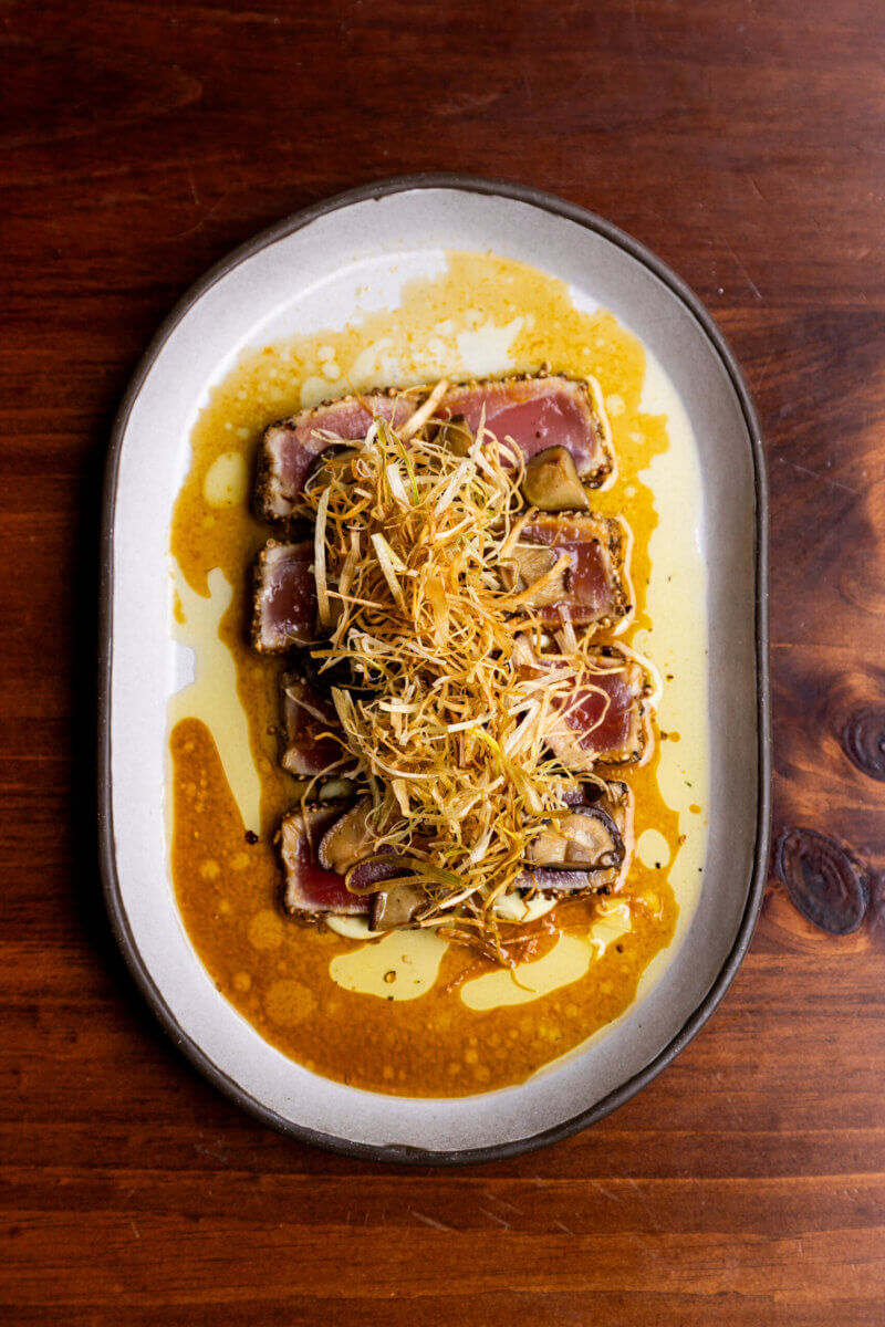 Six slices of seared tuna topped with shredded fried leeks, served on a white oval plate with a soy-based sauce, invite you to Eat Your Way Around the World—right in the heart of Birmingham. Presented on a wooden table, this dish offers an international flair in every bite.