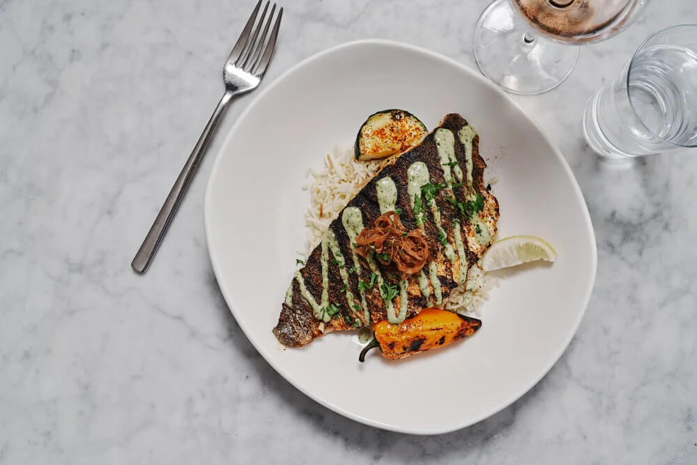 Grilled fish fillet with herb sauce served over rice, accompanied by grilled vegetables and a lime wedge, on a white plate with a fork, glass of water, and glass of wine nearby—an inviting meal that lets you Eat Your Way Around the World without leaving Birmingham.