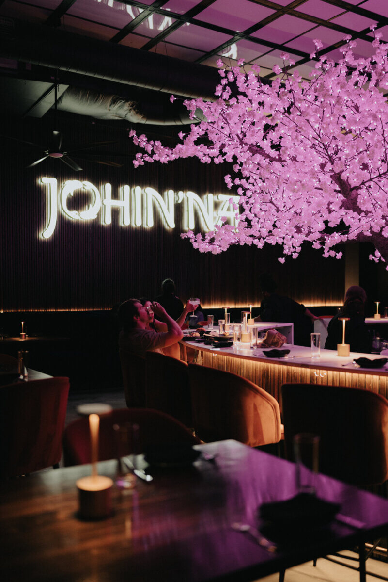 Southern Roots, Japanese Technique: Inside Chef Ashley Jackson's Johin'na - 6 Indoor restaurant scene with a large, lit faux cherry blossom tree and neon “JOHIN'NA” sign; guests dine and snap photos, savoring dishes by Chef Ashley.