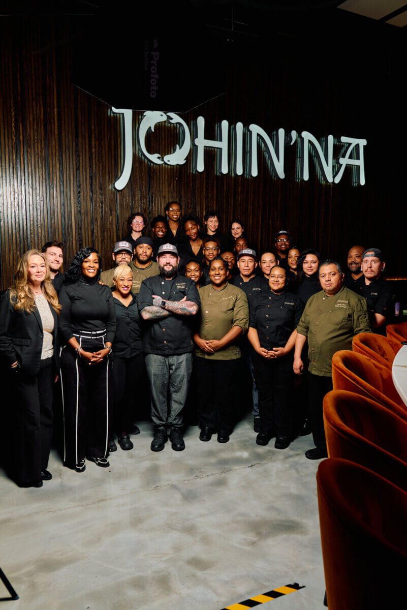 Southern Roots, Japanese Technique: Inside Chef Ashley Jackson's Johin'na - 5 A group of restaurant staff, including Chef Ashley, pose together in front of a dark wood wall with a “JOHIN’NA” sign overhead in Birmingham.