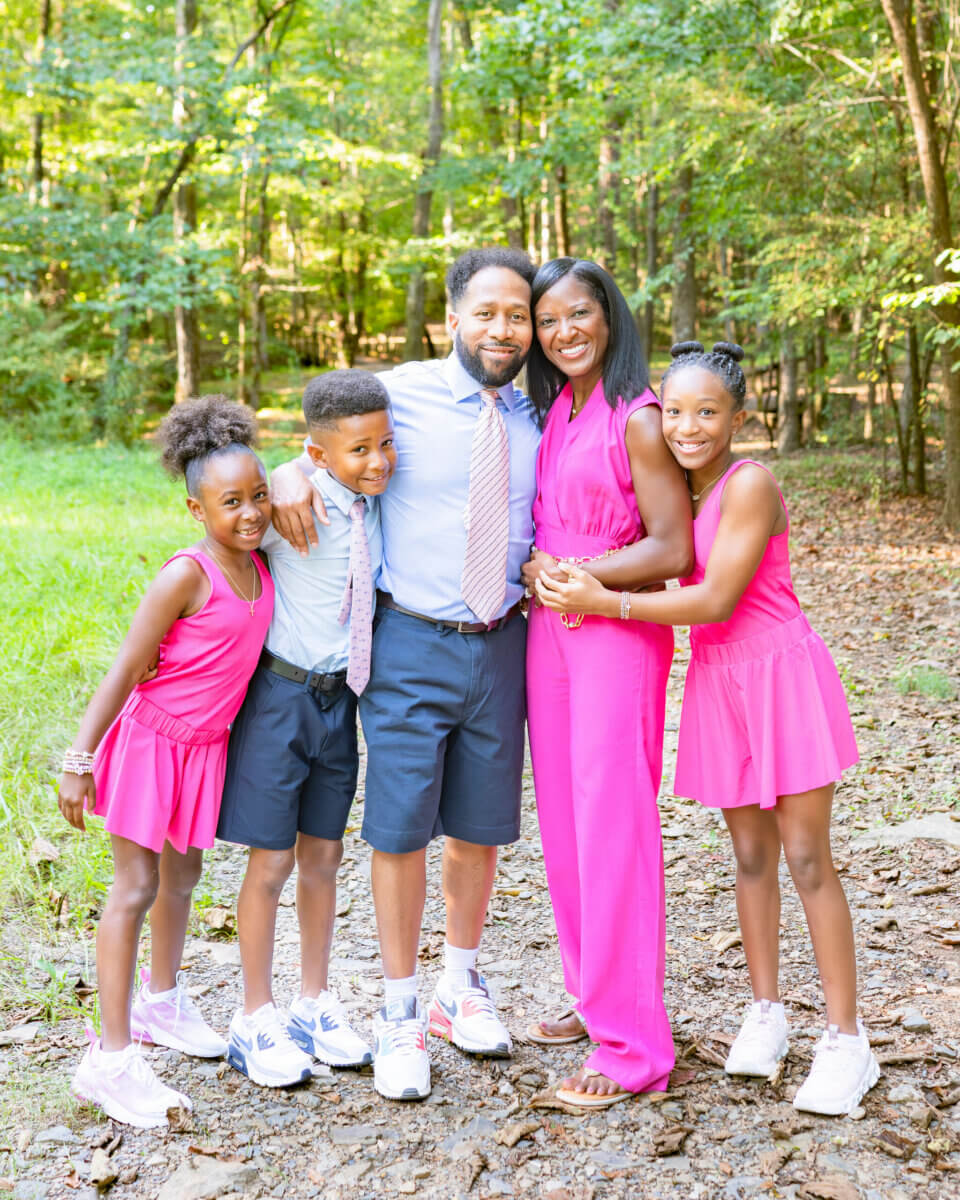 This BHAM Doctor Fights for Women's Lives Every Day - 2 A family of five stands together outdoors on a rocky path, surrounded by trees. The adults and three children smile at the camera; all are dressed in pink or blue attire, embodying the spirit of research and advocacy for women’s health.
