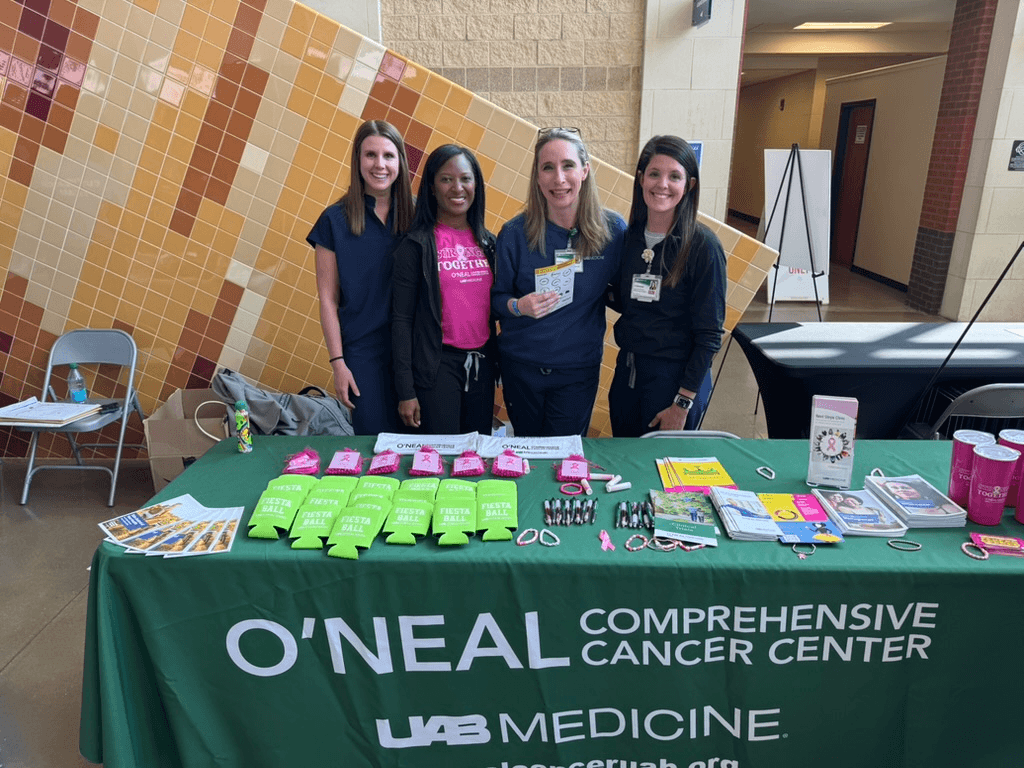 This BHAM Doctor Fights for Women's Lives Every Day - 3 Four women stand behind a table with informational materials and giveaways for O’Neal Comprehensive Cancer Center at a health event, highlighting their commitment to research and advocacy in improving women’s lives.