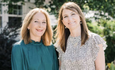 Two women with light brown hair are standing side by side outdoors, smiling at the camera. One wears a teal top while the other wears a white blouse with black spots, capturing a joyful moment that reflects their Family Legacy. Green foliage is in the background.