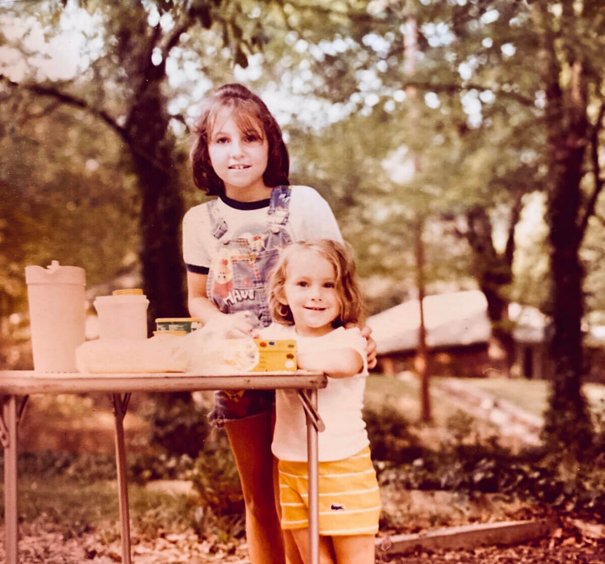 Two young children stand by a table with food and a pitcher outdoors, surrounded by trees and a house in the background, capturing the warmth of Gigi’s cherished family legacy.