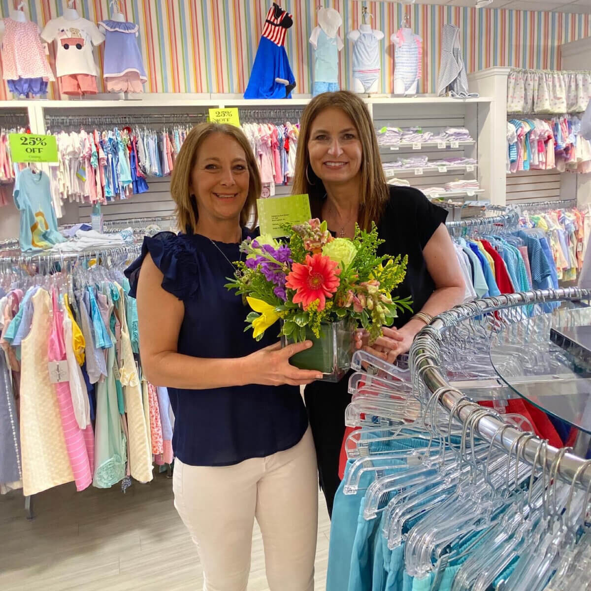Two women stand in Gigi’s children’s clothing store, holding a bouquet of flowers. Racks of colorful clothes and sale signs reflect the family legacy behind The Lili Pad, visible in the background.