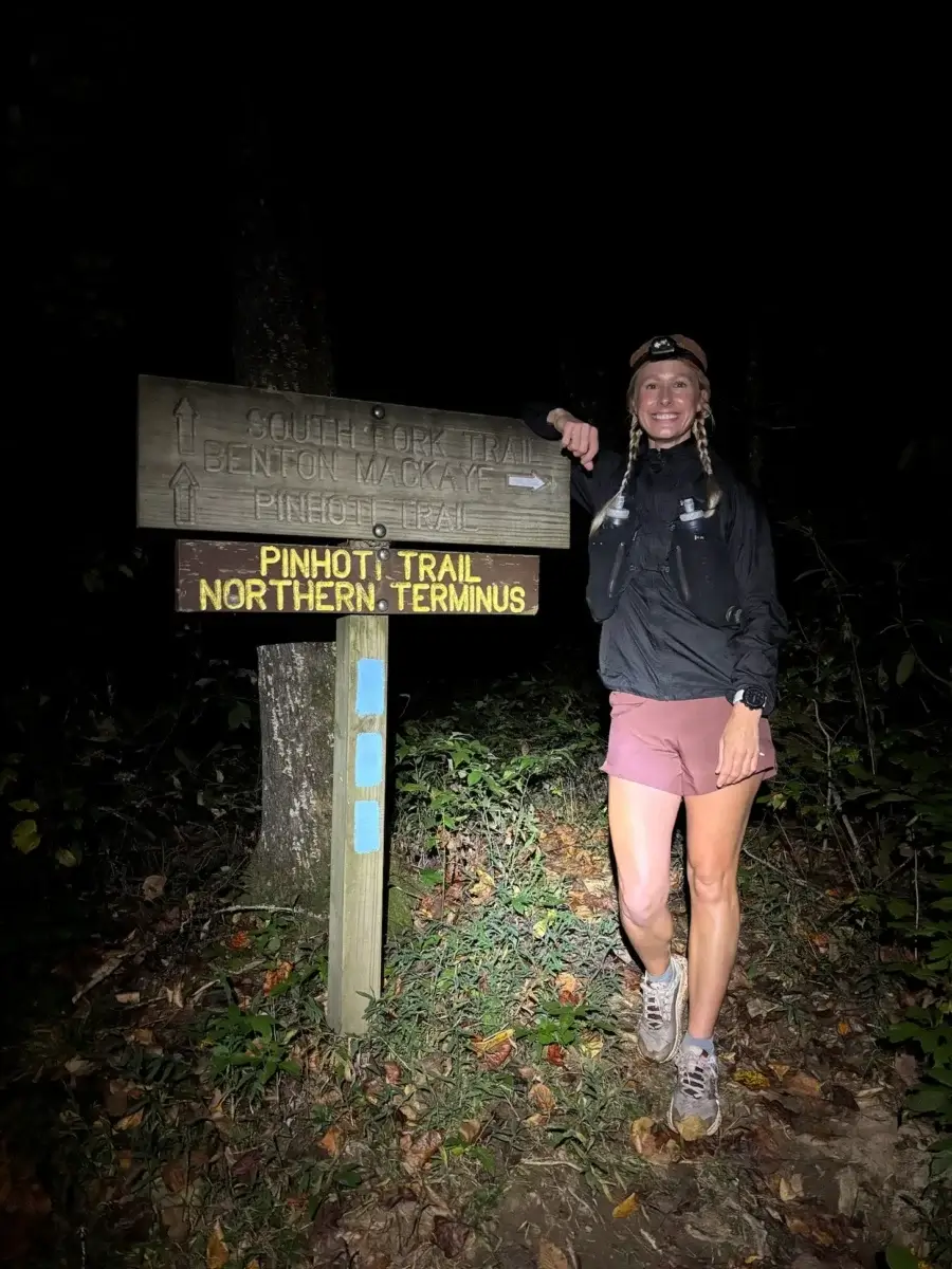 This BHAM Mom Ran 350 Miles in 5 Days - 3 A Birmingham woman stands beside a wooden trail sign at night, illuminated by a flashlight. The Pinhoti Trail Northern Terminus marks the start of her 350 miles in five days journey.