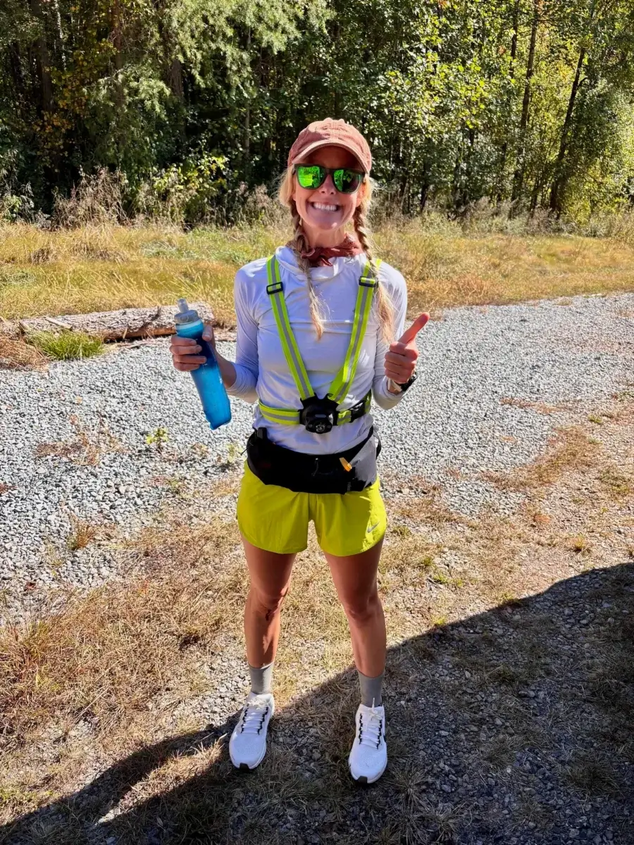 This BHAM Mom Ran 350 Miles in 5 Days - 2 Birmingham woman in running gear stands outdoors on a sunny day, holding a blue water bottle, giving a thumbs up after running 350 miles in five days. Trees and gravel path behind her.