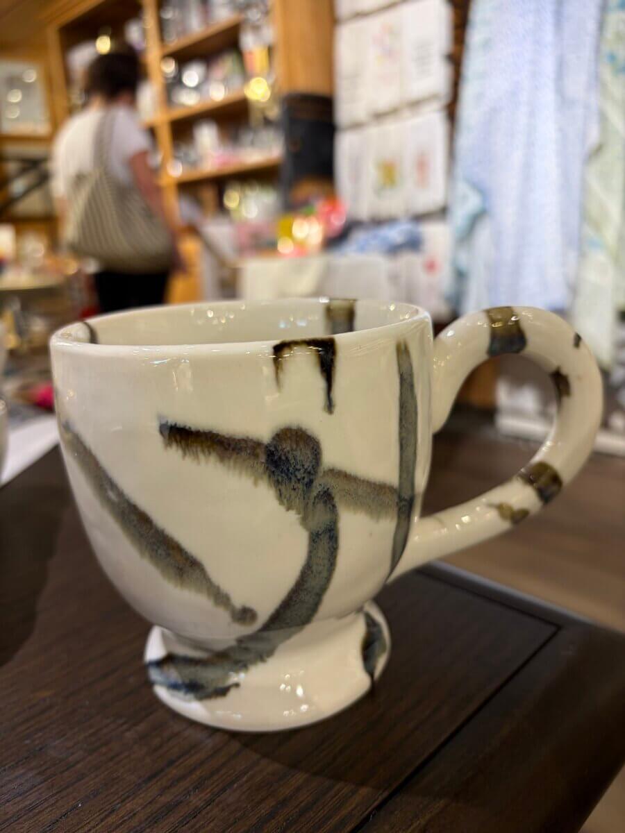 A ceramic mug with abstract blue and brown brushstrokes sits on a dark wooden table in a shop with shelves and a person in the background, making it a charming addition to your fall decor.