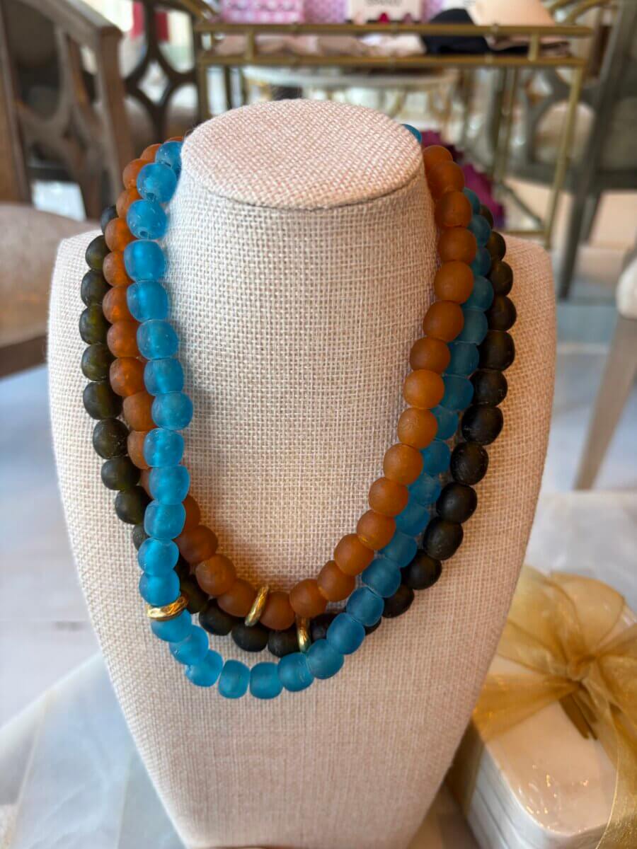 Three beaded necklaces—one turquoise, one amber, and one dark brown—are displayed on a beige necklace stand with a gold ring accent on the turquoise strand, making them perfect fall accessories to complement your seasonal style.