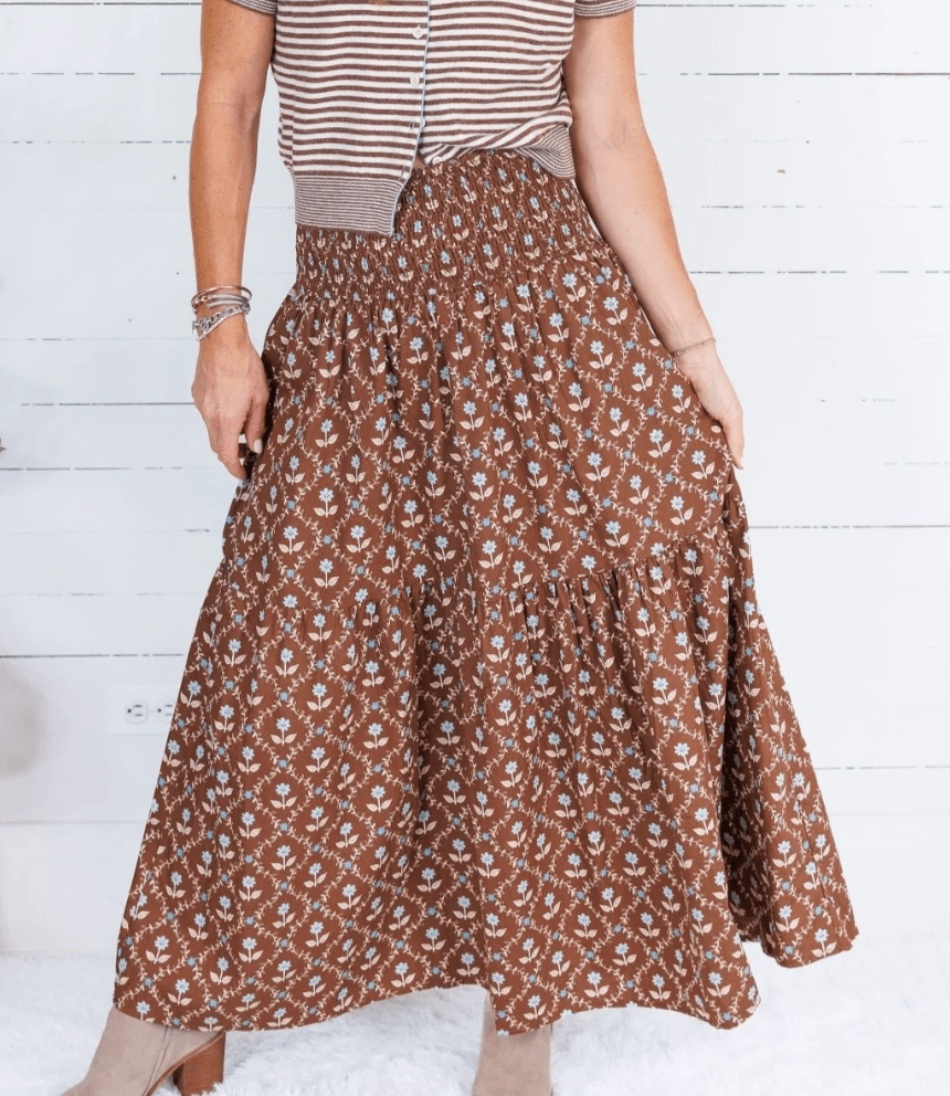 A person wearing a brown patterned maxi skirt, a short-sleeve striped top, and taupe boots—perfectly embodying fall fashion—stands in front of a white wall.