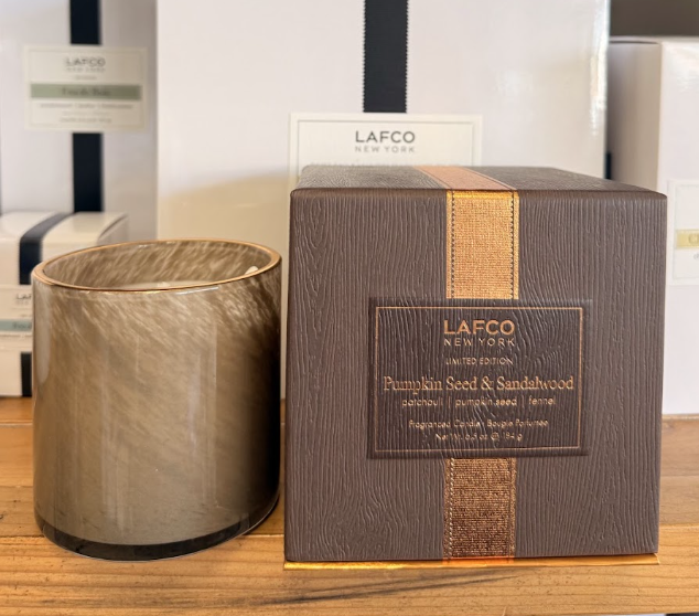 A LAFCO New York candle and its box labeled “Pumpkin Seed & Sandalwood” are displayed on a wooden surface in front of stacked white boxes, adding a touch of cozy fall decor to any space.