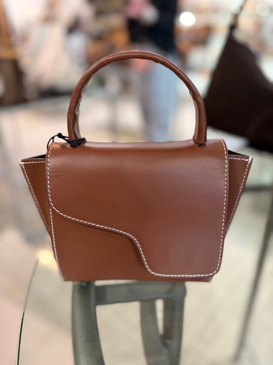 Brown leather handbag with white stitching sits on a glass surface, featuring a top handle and an asymmetrical front flap. Perfect for fall fashion, this stylish piece adds a sophisticated touch to your seasonal look. Background is blurred.