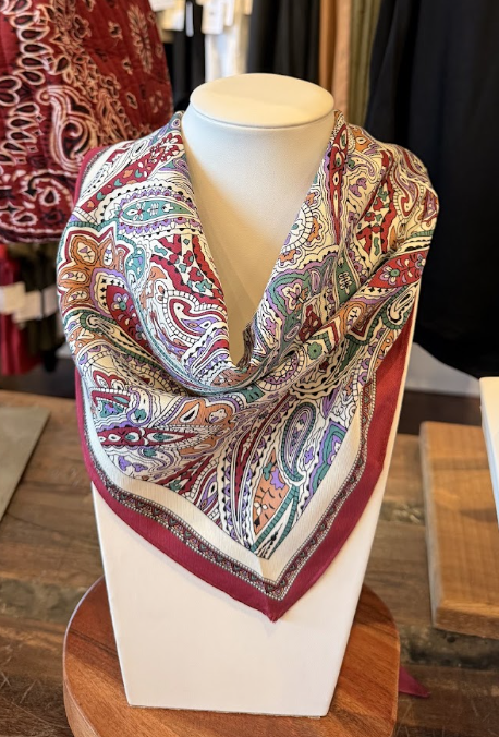 A mannequin displays a folded silk scarf with a colorful paisley pattern in red, green, and purple tones, bordered in dark red, on a wooden stand—an elegant addition to your fall accessories and seasonal wardrobe.