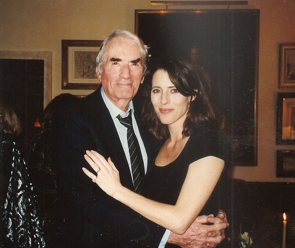 An older Gregory Peck in a suit and Cecilia Peck in a black dress stand close together, smiling at the camera.
