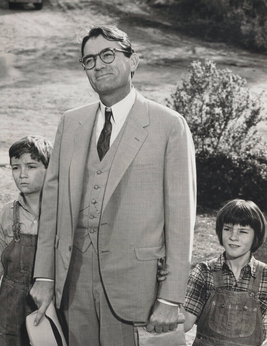 Atticus Finch in a suit stands outdoors holding hands his two children: Jem and Scout Finch, in the film To Kill a Mockingbird. 