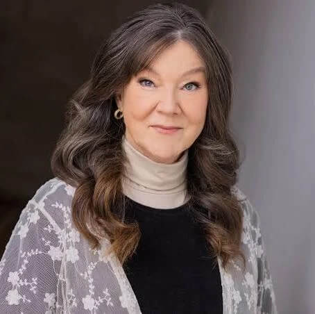 Mary Badham with long brown hair, wearing a white lace cardigan over a black top and beige turtleneck, poses in front of a neutral background.