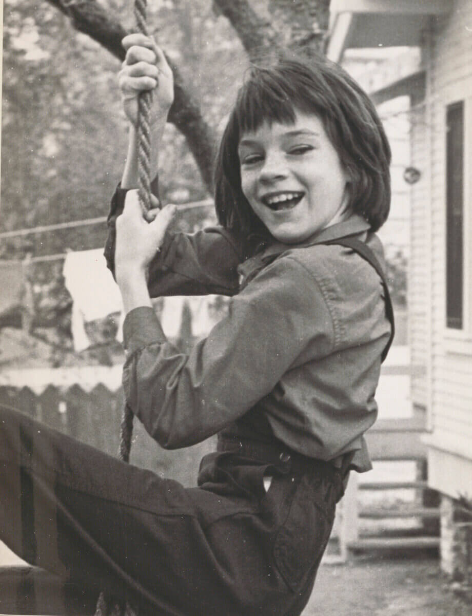 Mary Badham as Scout Finch with short hair and overalls smiles while holding onto a rope swing outdoors on the set of To Kill a Mockingbird.