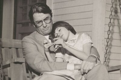 Gregory Peck as Atticus Finch in a suit sits on a porch swing, with Scout Finch played by Mary Badham resting in his lap. Image courtesy of Virginia Samford Theatre