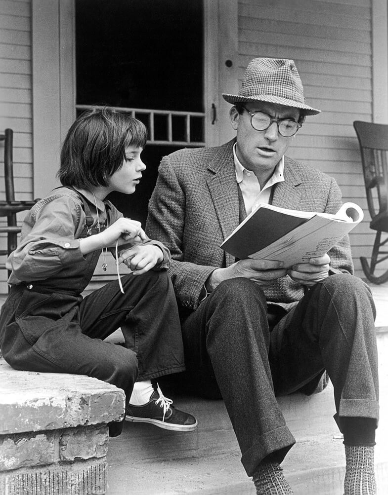 Gregory Peck wearing glasses and a hat reads from a book to Mary Badham sitting beside him on porch steps on the set of To Kill a Mockingbird.
