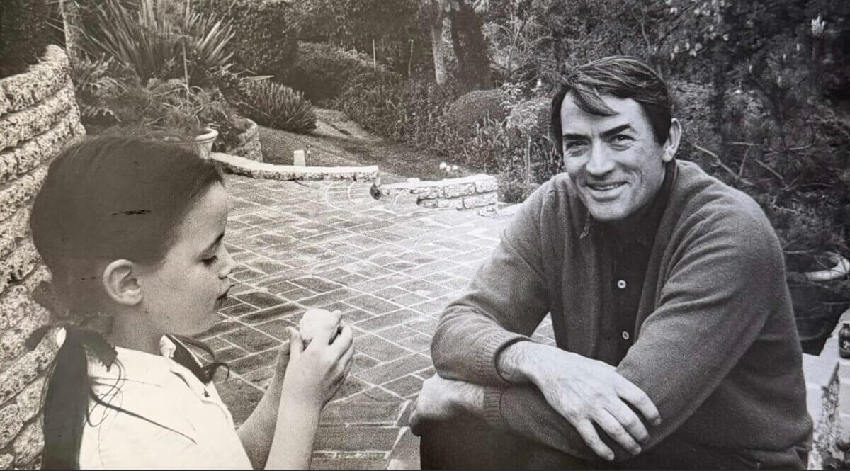 Gregory Peck on a brick patio, smiling at the camera, while Cecilia Peck stands nearby holding an apple.