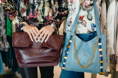 Two women stand side by side, one holding a large maroon ALC leather clutch, the other wearing a RAPT light blue shoulder bag with gold chain and metallic details. Both wear L'agence patterned tops—styled by Gus Mayer.