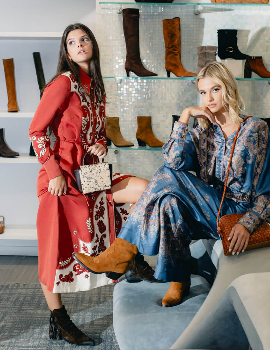 Minimalism Who? This Fall’s Trends Are All About Living Boldly Like Dolly - 9 Two women pose in a shoe store, showcasing Gus Mayer’s latest Fall Trends. They wear patterned long dresses and ankle boots with handbags, while shelves behind them display various pairs of boots.