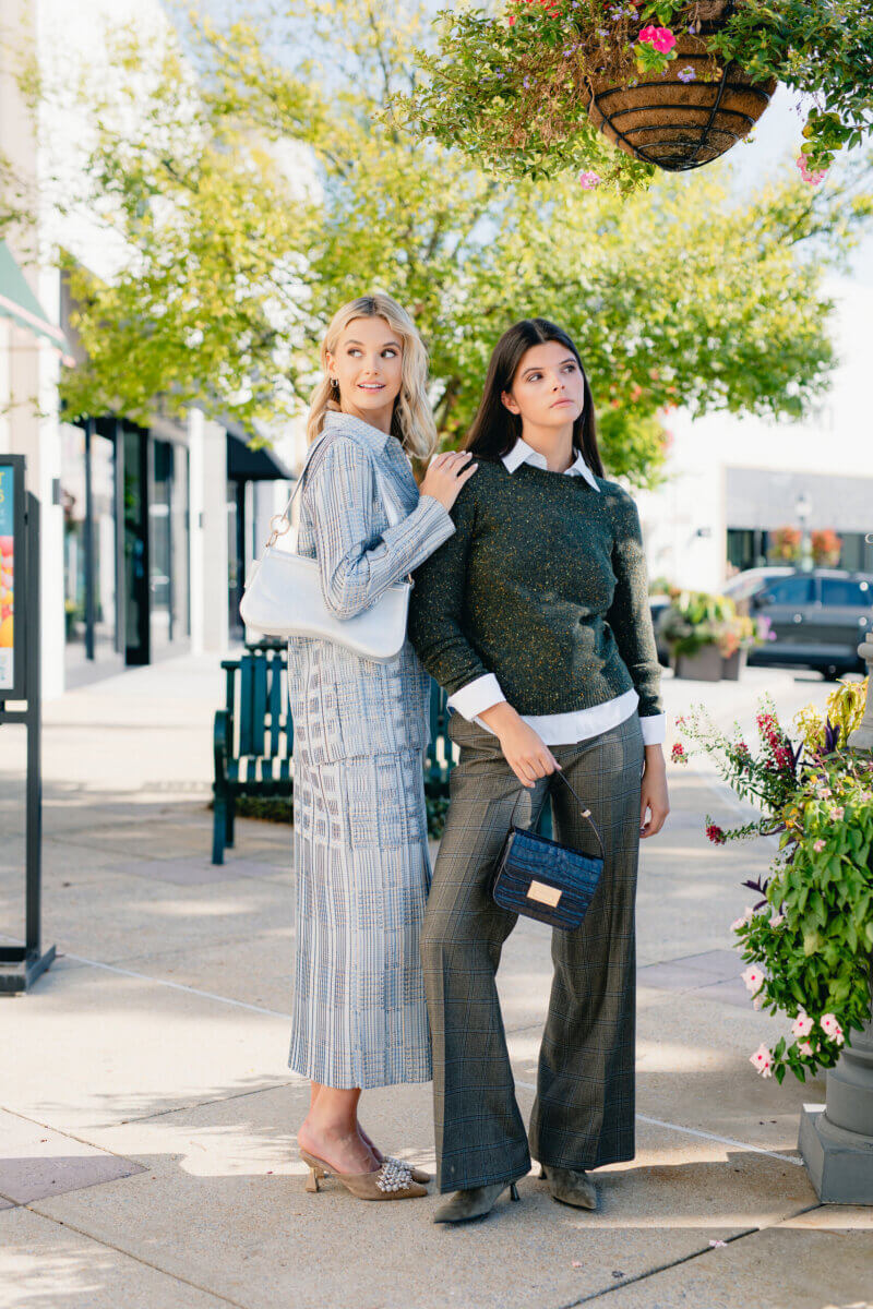 Minimalism Who? This Fall’s Trends Are All About Living Boldly Like Dolly - 4 Two women stand on a sidewalk, dressed in business casual outfits inspired by the latest Fall Trends from Gus Mayer, with greenery and flowers around them and shops in the background on a sunny day.