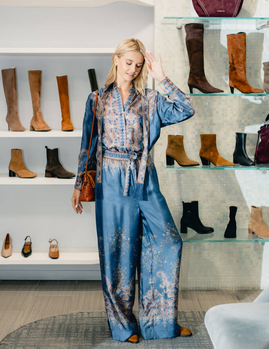 Minimalism Who? This Fall’s Trends Are All About Living Boldly Like Dolly - 10 A woman in a silky blue patterned outfit stands in Gus Mayer, surrounded by shelves displaying various boots and heels—perfect inspiration for your Fall Trends lookbook.