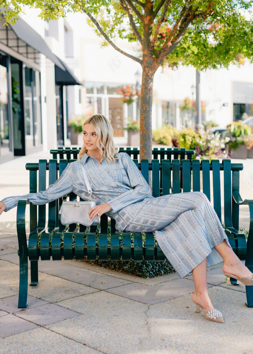 Minimalism Who? This Fall’s Trends Are All About Living Boldly Like Dolly - 5 Woman in a blue plaid dress sits on a green bench under a tree, holding a white purse and wearing heeled sandals in an outdoor shopping area—capturing effortless style perfect for the Gus Mayer Fall Trends Guide.