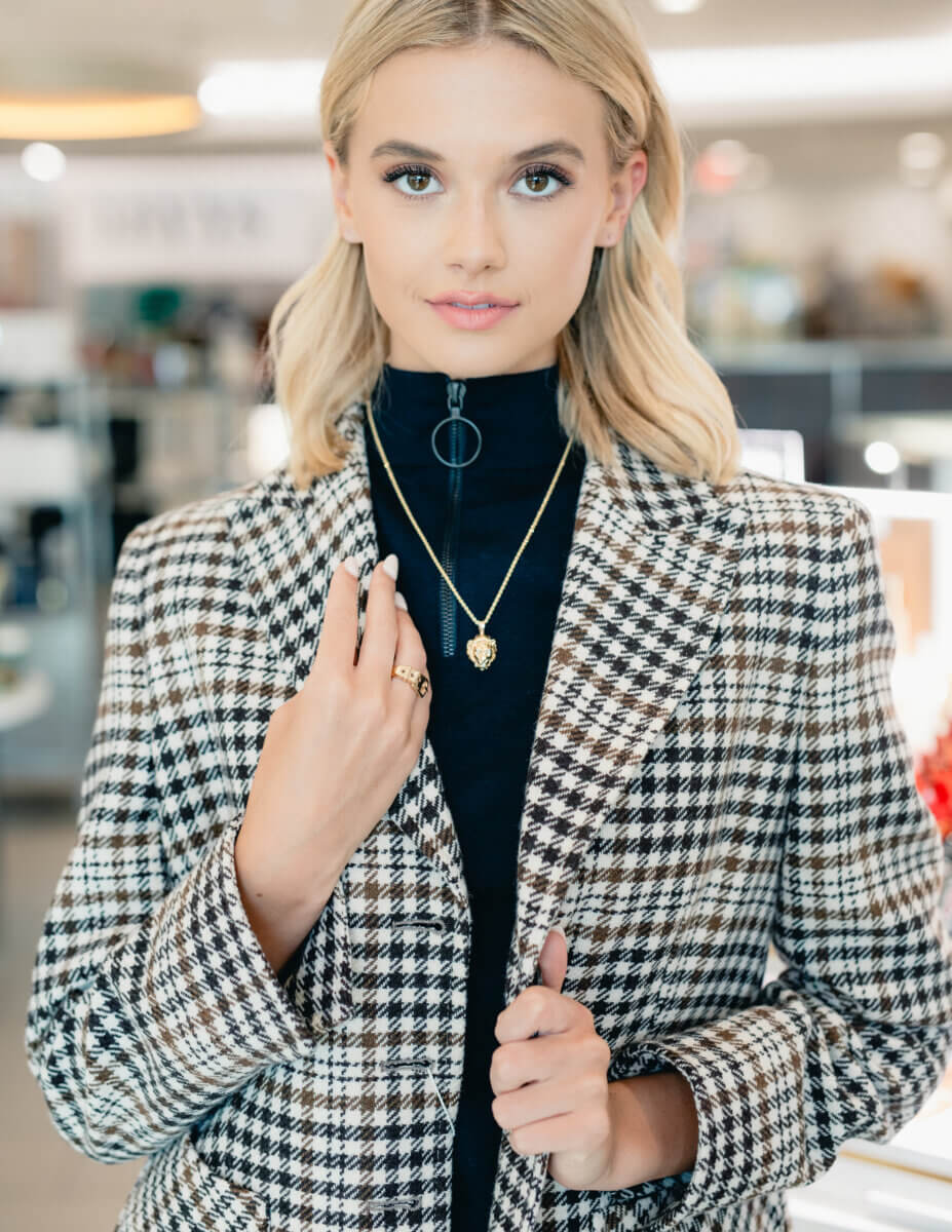 Minimalism Who? This Fall’s Trends Are All About Living Boldly Like Dolly - 8 A woman with blonde hair wears a black turtleneck, a houndstooth blazer, and gold jewelry—showcasing one of the top Fall Trends from Gus Mayer—while standing indoors, looking directly at the camera.