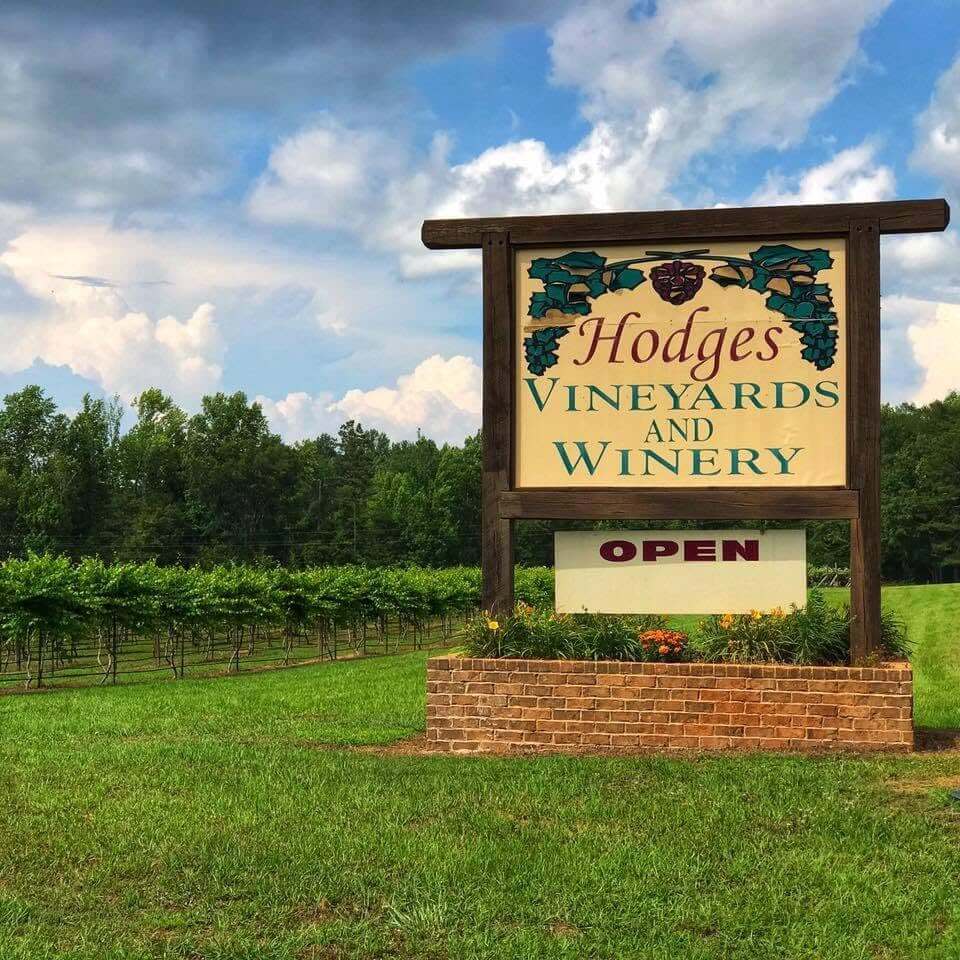 The large wooden sign for Hodges Vineyards and Winery stands by rows of grapevines, with a grassy field and trees in the background under a cloudy sky, making it one of the gorgeous wineries near Birmingham.