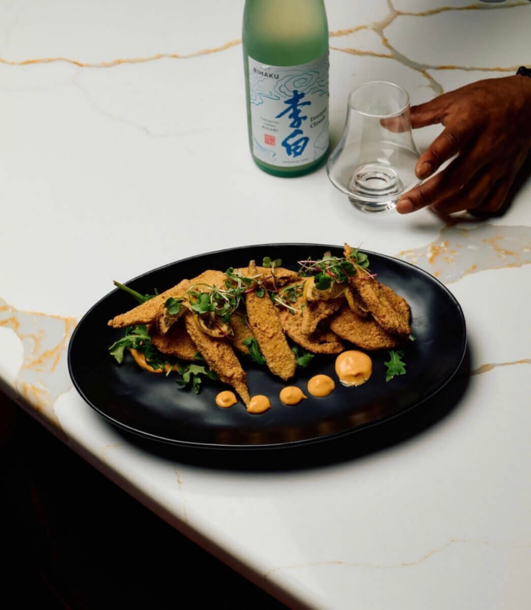 A black plate with breaded fried slices, garnished with greens and sauce dots, invites you to Eat Your Way Around the World. A bottle of sake, a glass, and a hand appear on a white marble table—bringing a taste of international flavor right to the heart of Birmingham.