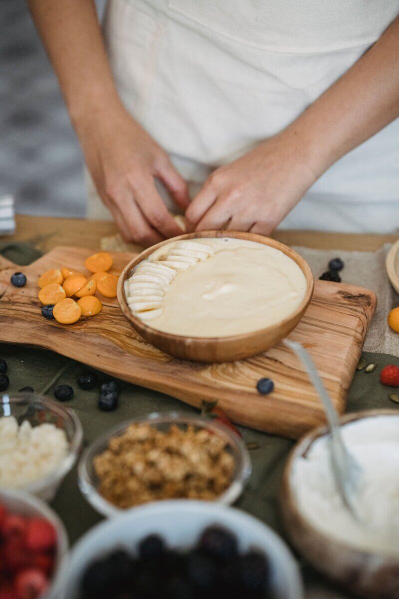 Pumpkin Is Not Just for Lattes — Supercharge Your Fall Smoothies and Salads! - 3 Person arranging fruit slices on a bowl of creamy dessert, with various fruits, granola, and yogurt—perfect for salads or fall smoothies—set out on a wooden board.