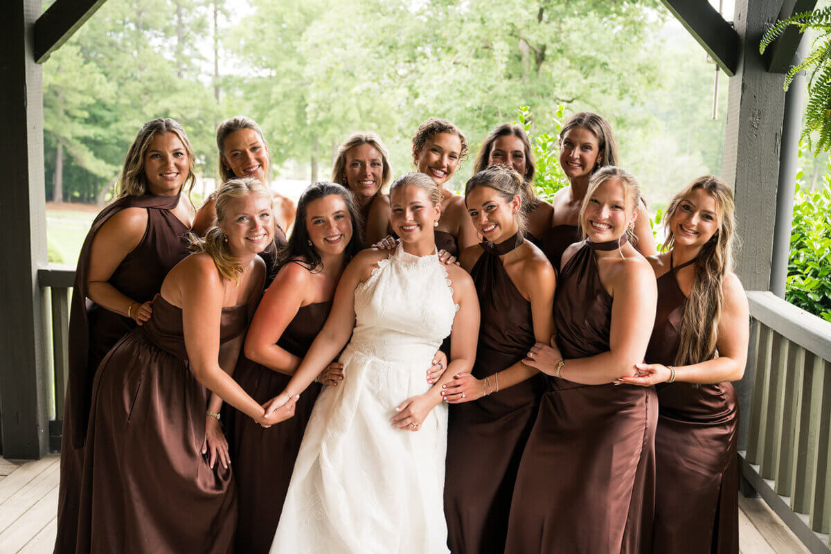 A bride in a white dress stands with eleven bridesmaids in brown dresses, all smiling and posing together on a covered porch with greenery in the background, capturing the joyful spirit of their colorful summer wedding.