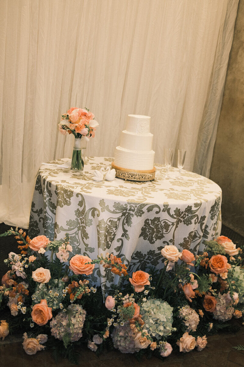 A three-tier white wedding cake on a decorated table with a floral arrangement, a bouquet in a vase, two champagne flutes, and a patterned tablecloth in front of a draped backdrop perfectly captures the spirit of a Colorful Summer Wedding, highlighted by vibrant blooms and thoughtful personal touches throughout the elegant display.