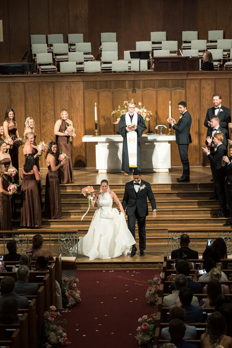 A bride and groom walk down the aisle in a church as wedding guests and attendants stand and applaud, celebrating a colorful summer wedding filled with personal touches.