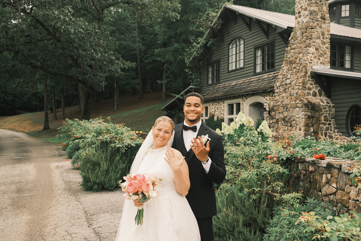 A bride and groom stand outside a rustic stone house, smiling and showing their wedding rings. The bride holds a bouquet of pink flowers, adding personal touches to their special day, and both are dressed in formal wedding attire perfect for a colorful summer wedding.