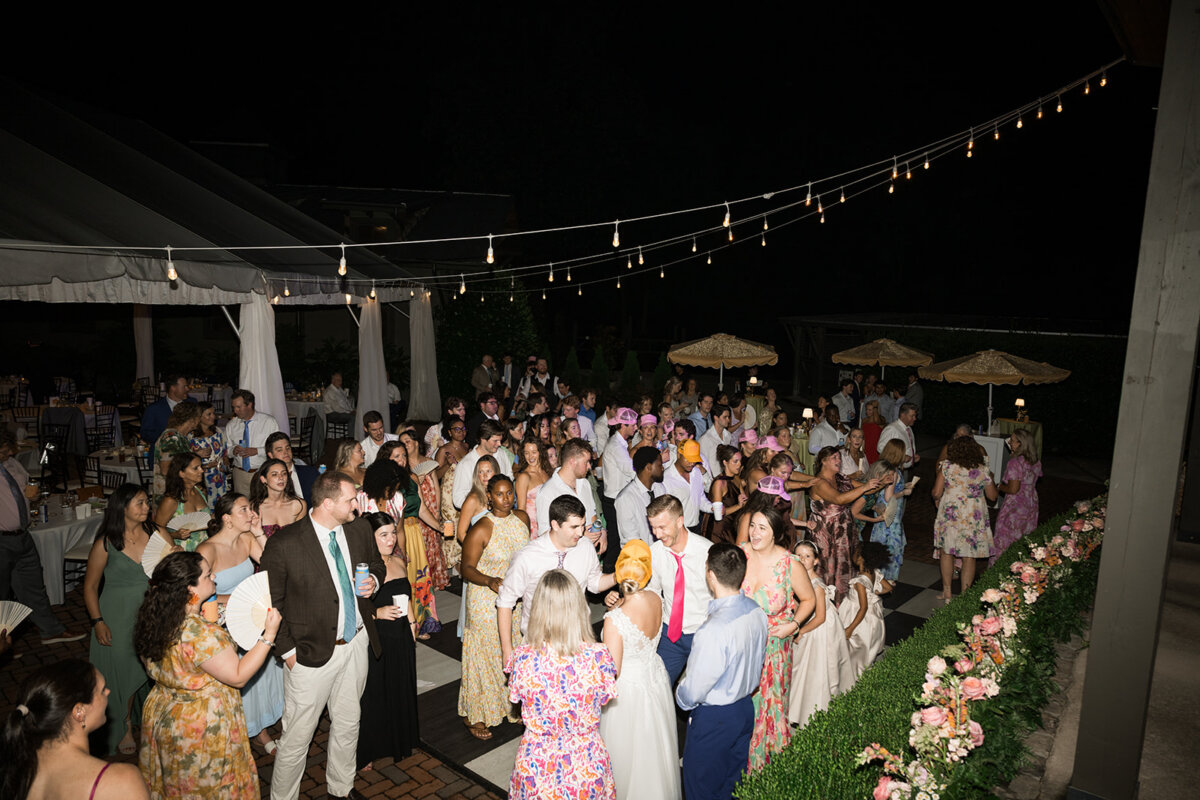 A large group of people dressed in formal and semi-formal attire gather and socialize at an outdoor nighttime event under string lights, enjoying the vibrant ambiance of a colorful summer wedding filled with charming personal touches.